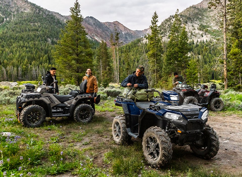 Multiple Polaris Sportsman ATVs parked at a mountain campsite, with riders preparing gear in a forested alpine setting.