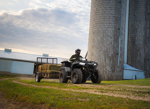 Polaris Sportsman ATV towing a trailer loaded with hay across a farm, demonstrating utility and work capabilities near barns and silos.