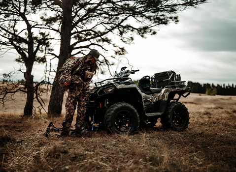 Camouflage Polaris Sportsman ATV parked in a field beside a hunter with archery equipment, highlighting hunting and outdoor adventure capabilities.