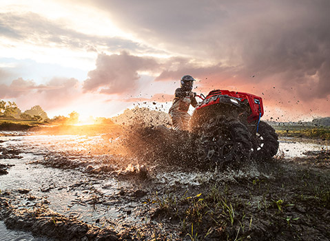 Polaris Sportsman XP 1000 High Lifter Edition ATV powering through a muddy water hole, spraying mud and water during an off-road mudding ride at sunset.
