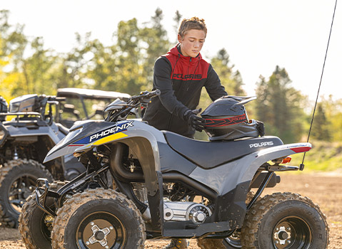 Polaris Phoenix youth ATV parked outdoors as a young rider places a helmet on the seat, with a Polaris Sportsman ATV visible in the background.