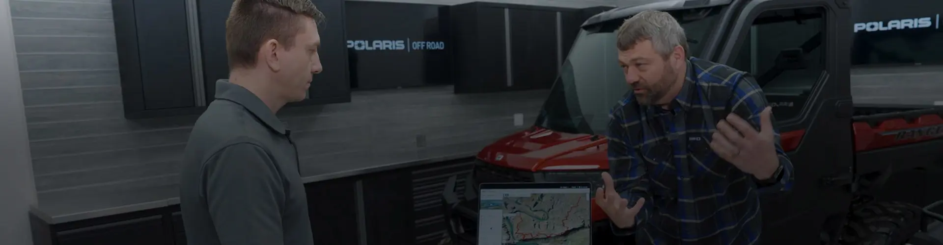 Pat McArdle and his friend Matt review Polaris RIDE COMMAND mapping features on a laptop inside a Polaris Off Road showroom, standing beside a red Polaris RANGER UTV.