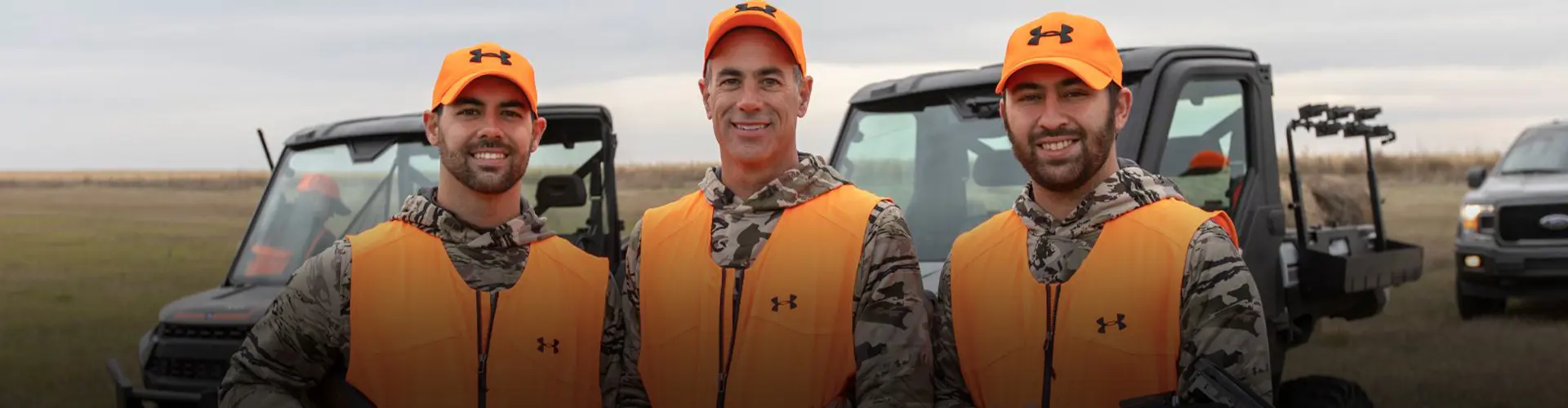 Three participants in blaze-orange hunting gear standing in a field with Polaris RANGER XP 1000 UTVs during the Ultimate Hunt Experience.