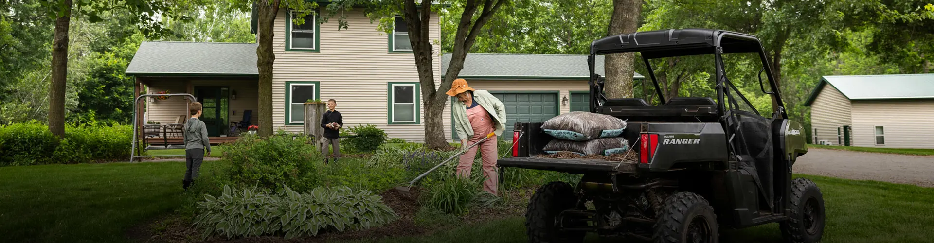 A black Polaris RANGER UTV parked in a backyard with its cargo bed loaded with bags of mulch. A woman is gardening with a rake, while two children are in the background.