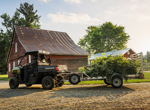 A Polaris RANGER UTV in green is shown pulling a trailer full of tree branches and yard waste on a gravel driveway in front of a rustic red barn.