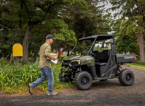 A man carrying a package and mail walks past a military green Polaris RANGER UTV parked on a gravel driveway next to a yellow mailbox.