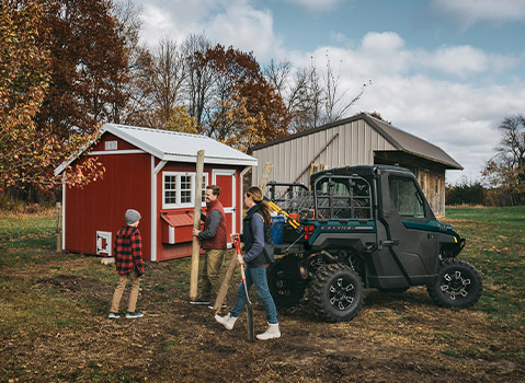 A family with a child carrying lumber near a newly built red chicken coop, with a dark green Polaris RANGER UTV parked nearby.