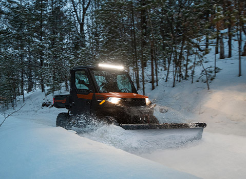 An orange and black Polaris RANGER UTV equipped with a snow plow and LED light bar, actively clearing a snowy path through a dense winter forest.