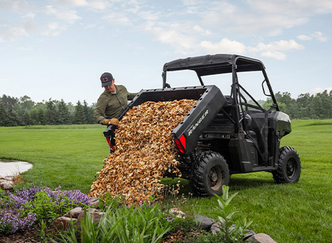 A man in work gloves is tilting the cargo bed of a black Polaris RANGER UTV, dumping a large pile of wood chips into a vibrant flower garden.