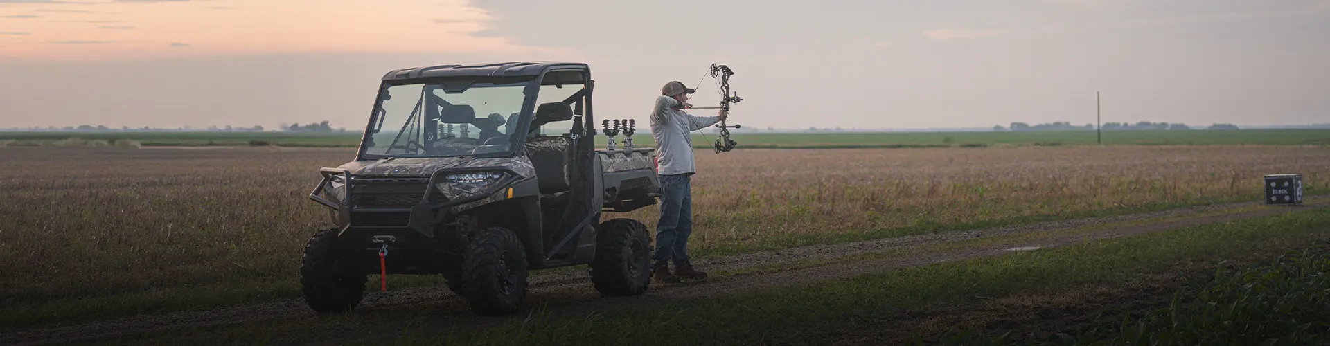 Luke Brewster aiming a compound bow beside a Polaris RANGER UTV on a farm trail at sunset, surrounded by open fields.