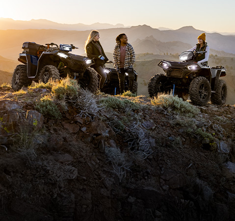 Three friends standing with two Polaris Sportsman 4-Wheeler ATVs on a scenic mountain ridge at sunset.
