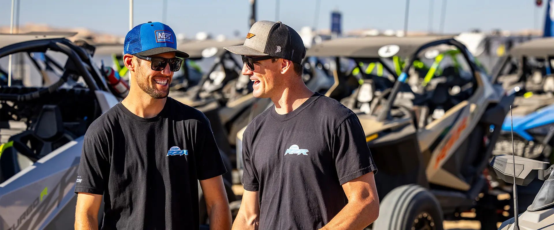 Two Camp RZR attendees stand in front of a lineup of Polaris RZR off-road vehicles.
