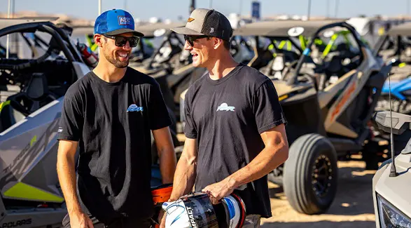 Two Camp RZR attendees stand in front of a lineup of Polaris RZR off-road vehicles.