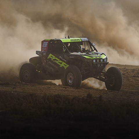 Black and green Polaris RZR Pro R Factory racing SxS speeding across desert terrain and kicking up a large cloud of dust.