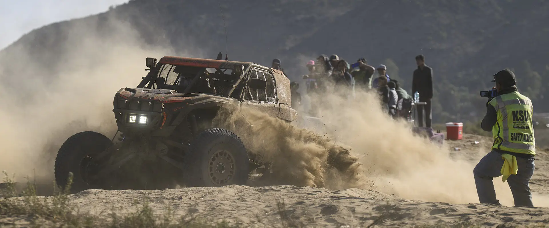 Mud-covered Polaris RZR racing SxS powering through sandy terrain as spectators and event staff watch.
