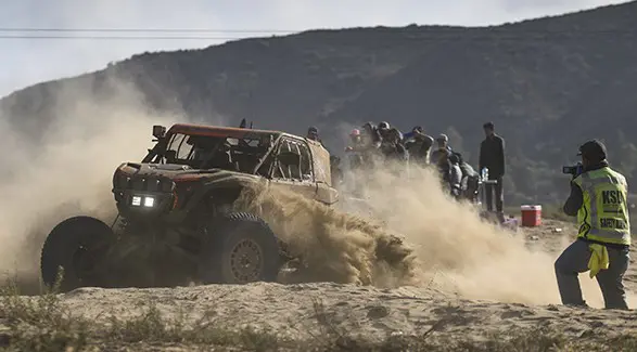 Mud-covered Polaris RZR racing SxS powering through sandy terrain as spectators and event staff watch.