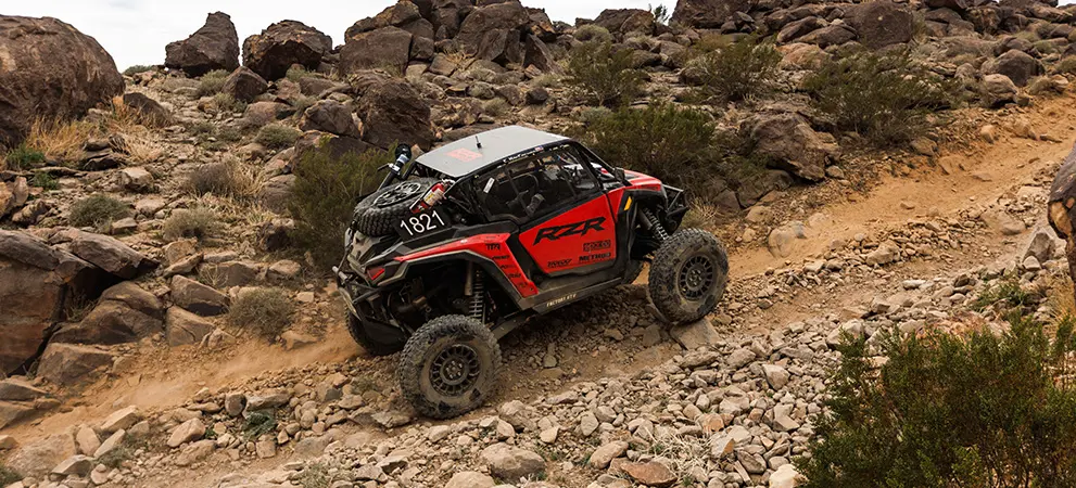 Polaris RZR Pro R Factory Side-by-Side climbing a steep rocky trail in desert terrain, kicking up dust among scattered boulders and brush.