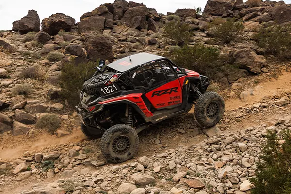 Polaris RZR Pro R Factory Side-by-Side climbing a steep rocky trail in desert terrain, kicking up dust among scattered boulders and brush.