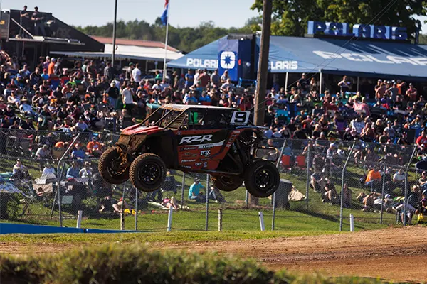 Polaris RZR Pro R Factory Side-by-Side airborne over a dirt jump during an off-road race, with a large crowd and Polaris tents in the background.