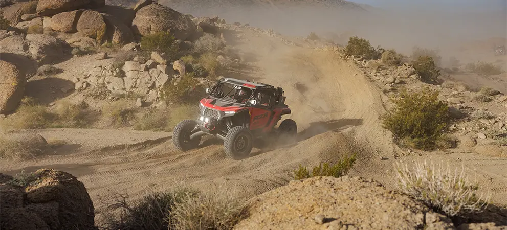 Polaris RZR Pro R Factory Side-by-Side racing through a dusty desert trail, carving a turn between rocks and low vegetation.