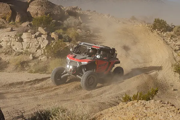 Polaris RZR Pro R Factory Side-by-Side racing through a dusty desert trail, carving a turn between rocks and low vegetation.