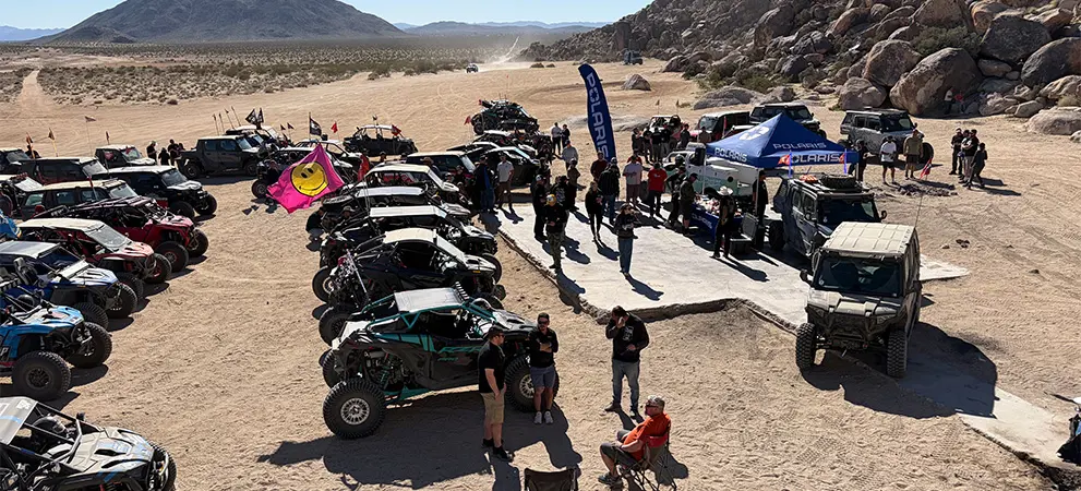 Large gathering of Polaris RZR Side-by-Side vehicles parked in a desert meetup, with attendees walking between rows near a central event setup.