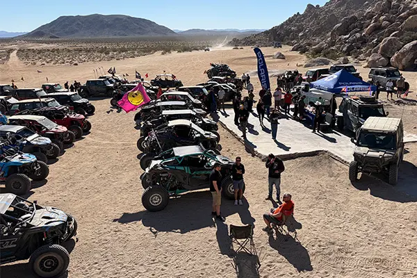 Large gathering of Polaris RZR Side-by-Side vehicles parked in a desert meetup, with attendees walking between rows near a central event setup.