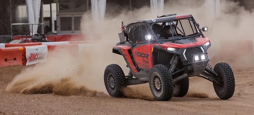 Polaris RZR Pro R Factory Side-by-Side drifting through a dirt corner during an off-road race, throwing dust with suspension extended and lights illuminated.