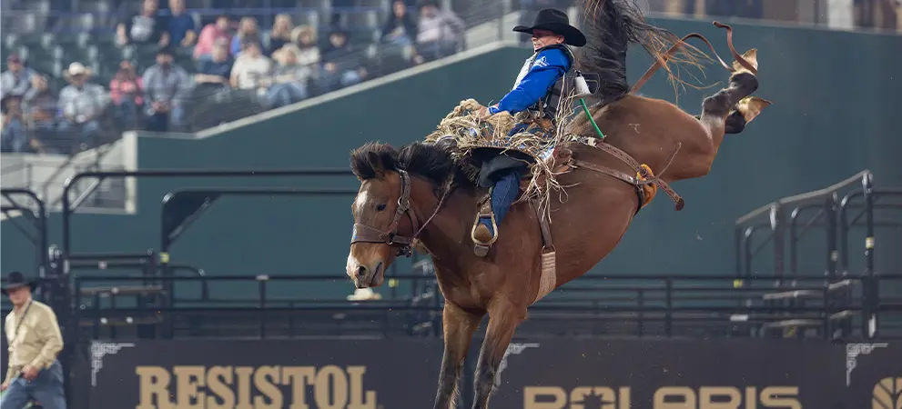 Bronco rider mid-jump on a bucking horse inside a rodeo arena, with dirt flying and sponsor signage visible in the background.