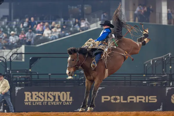 Bronco rider mid-jump on a bucking horse inside a rodeo arena, with dirt flying and sponsor signage visible in the background.