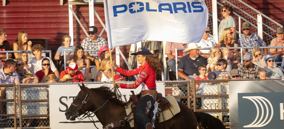 Rider on horseback carrying a Polaris flag along a rodeo arena fence, passing a crowded grandstand under bright daylight.
