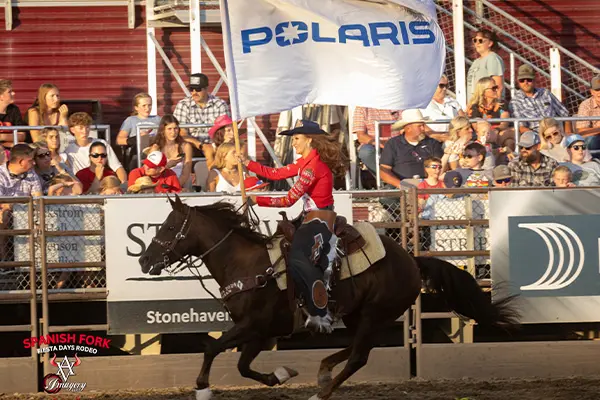 Rider on horseback carrying a Polaris flag along a rodeo arena fence, passing a crowded grandstand under bright daylight.