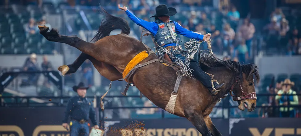 Bronco rider in motion on a bucking horse in a rodeo arena, surrounded by blurred spectators and arena barriers.