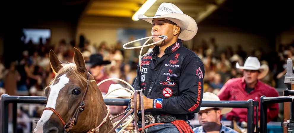 Rider on horseback holding a lasso in mouth while preparing to rope at a rodeo event, with other participants nearby.