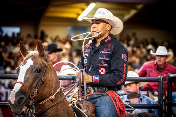 Rider on horseback holding a lasso in mouth while preparing to rope at a rodeo event, with other participants nearby.