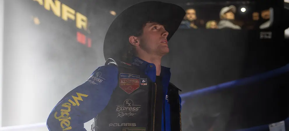 Rodeo competitor standing under arena lighting wearing protective gear and sponsor patches, with a dark, smoky background.