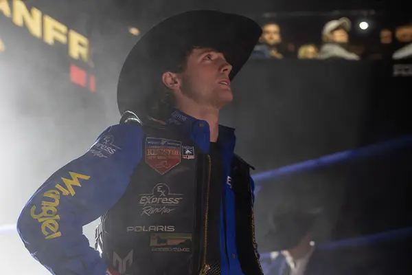 Rodeo competitor standing under arena lighting wearing protective gear and sponsor patches, with a dark, smoky background.
