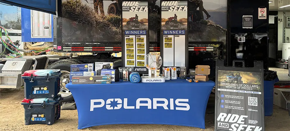Polaris branded table displaying gear and prizes for a Ride and Seek promotion, set up in front of a trailer with signage and equipment.