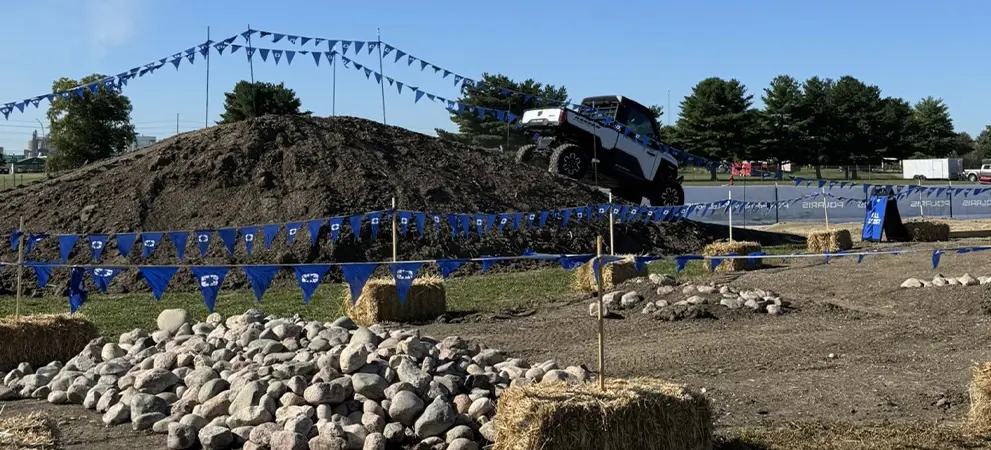 Polaris RANGER UTV climbing a dirt mound on an off-road demo course marked with blue flags, rocks, and hay bales under a clear sky.