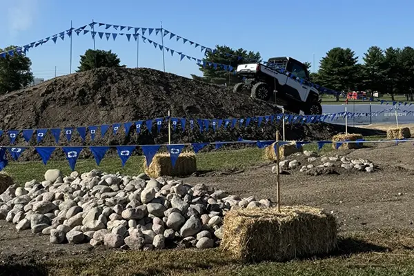 Polaris RANGER UTV climbing a dirt mound on an off-road demo course marked with blue flags, rocks, and hay bales under a clear sky.