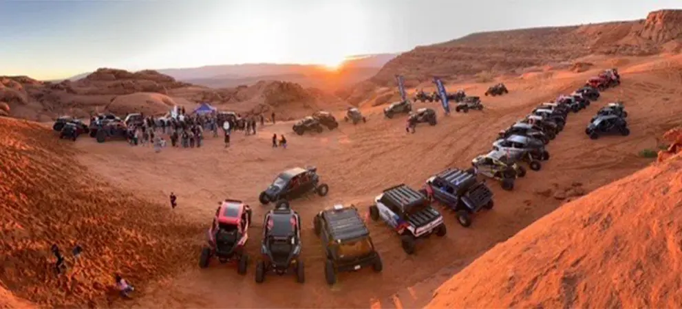 Large group of Polaris RZR Side-by-Side vehicles gathered in a desert valley at sunset, with participants and off-road vehicles lined across red sand terrain.