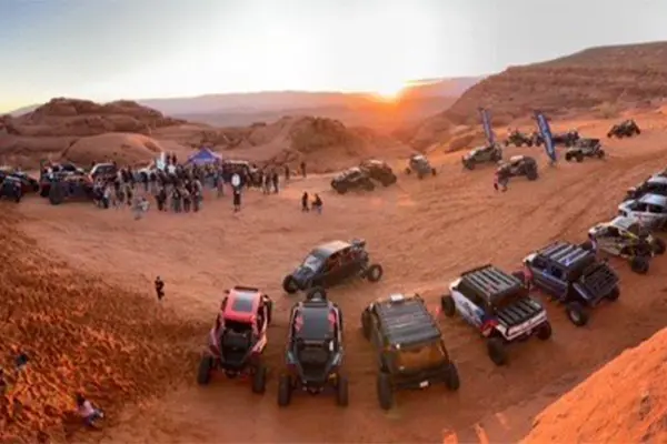 Large group of Polaris RZR Side-by-Side vehicles gathered in a desert valley at sunset, with participants and off-road vehicles lined across red sand terrain.