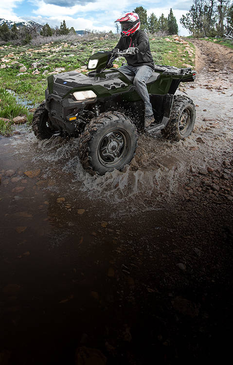 Polaris Sportsman ATV rider crossing a shallow stream on a mountain trail, surrounded by grassy hills, rocky terrain, leafless trees, and snow-capped peaks under a cloudy sky.