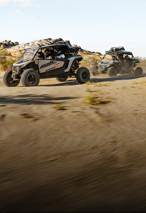 Two Polaris RZR XP S 4 Ultimate vehicles driving across a dusty desert trail with rocky hills in the background.