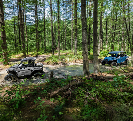 A white Polaris GENERAL XP 1000 and a blue Polaris GENERAL XP 4 1000 navigate a muddy forest trail and water crossing.