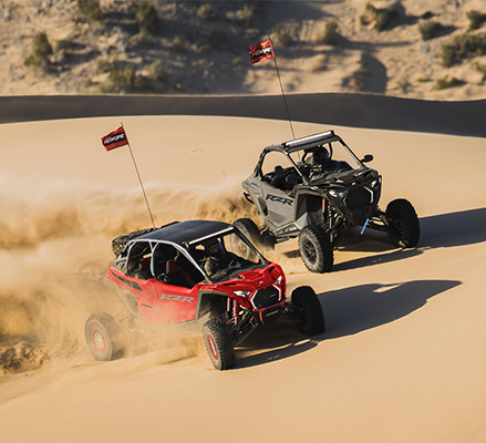 A red Polaris RZR Pro R 4 Ultimate and a grey Polaris RZR Pro S carve through high desert sand dunes, kicking up a large plume of sand.