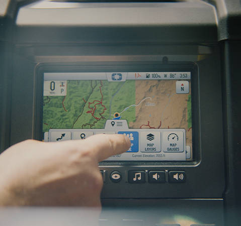 A hand interacts with the Polaris RIDE COMMAND touchscreen to select Group Ride features, showing a detailed off-road trail map and integrated vehicle gauges.