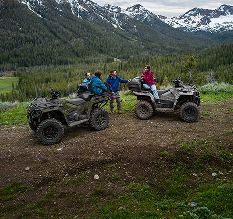 Two Polaris Sportsman ATV models parked on a mountain overlook trail, with four people gathered nearby and a forested valley stretching across the background.