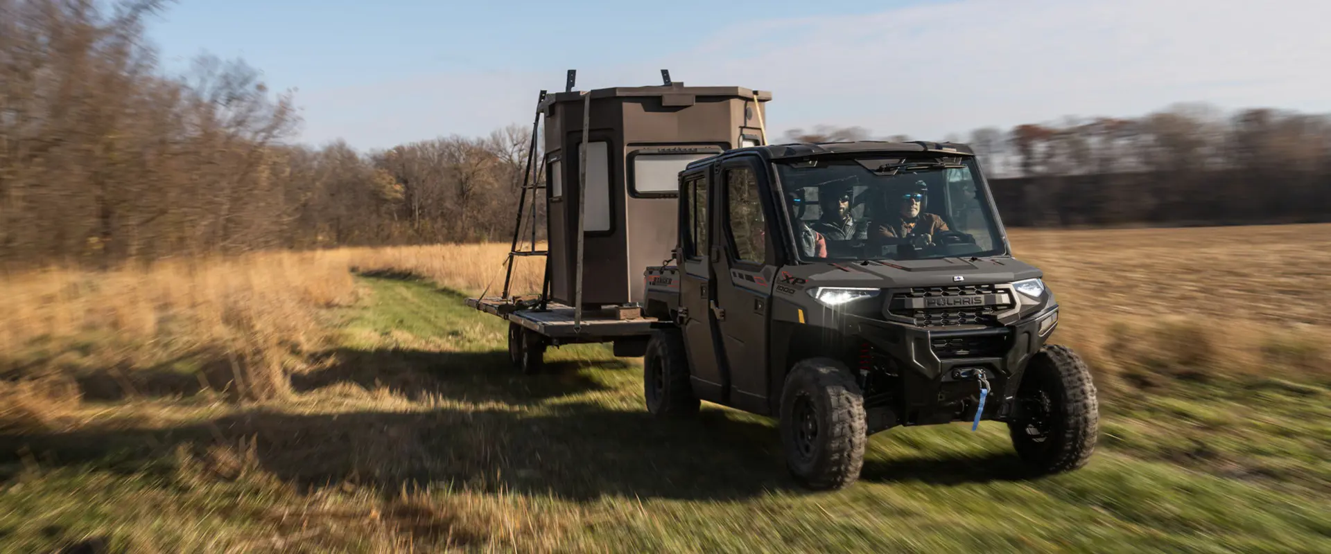 A 2026 Polaris RANGER CREW XP 1000 NorthStar Edition UTV pulling a trailer with a hunting shack on it.