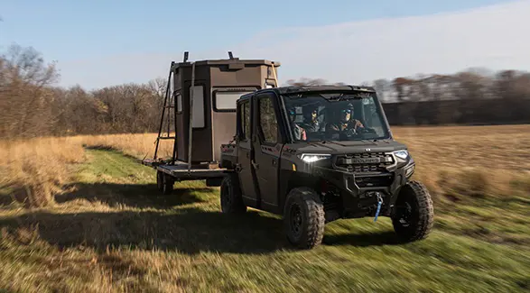 A 2026 Polaris RANGER CREW XP 1000 NorthStar Edition UTV pulling a trailer with a hunting shack on it.